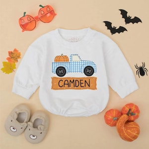 May include: White long-sleeved baby bodysuit with a blue gingham truck carrying pumpkins. The name "CAMDEN" is printed on a brown banner below the truck. Halloween-themed decorations surround the bodysuit.