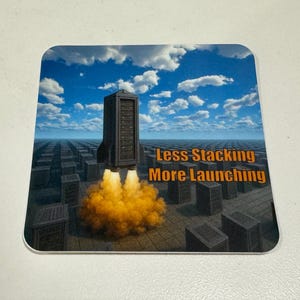 May include: Square coaster with a surreal image of a server tower launching like a rocket, with a blue sky and clouds background. The text "Less Stacking More Launching" is displayed on the coaster.