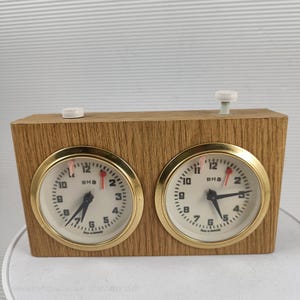 May include: A vintage chess clock with a wood grain finish. The clock features two round faces with gold-colored bezels, black hands, and the text "BHB". The clock has a white button on top and is marked "Made in Germany".