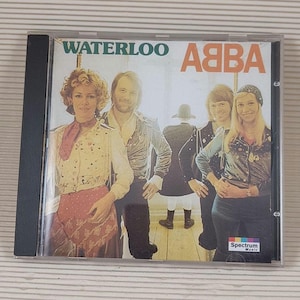 Op de afbeelding: Een CD-hoes met een gele en witte achtergrond waarop een band van vier personen te zien is. De band heet ABBA en het album heet Waterloo. De albumhoes toont de bandleden in verschillende poses, waaronder staand, zittend en leunend tegen een muur. De albumhoes toont ook een logo voor Spectrum Music.
