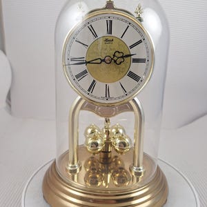 Peut inclure: Une horloge anniversaire à quartz dorée sous un dôme en verre transparent. Elle présente un cadran blanc avec des chiffres romains noirs, un centre doré avec des détails en filigrane et quatre boules de pendule rotatives sur une base ronde.