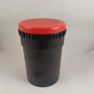 Może przedstawiać: Czarny zbiornik do wywoływania filmów z czerwoną pokrywką. Cylindryczny pojemnik ma żebrowaną krawędź u góry i gładki korpus. Pokrywka jest w jednolitym, jasnoczerwonym kolorze.