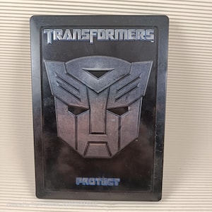 Op de afbeelding: Een zwarte metalen koffer met het Transformers-logo en het woord "PROTECT" in blauwe tekst.