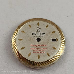 May include: A vintage watch face with a gold-colored, ridged outer ring. The face is off-white with gold-colored markers and the brand name "MORTIMA". Text includes "Super Automatic", "Unbreakable Spring", "Shockproof", and "Antimagnetic".