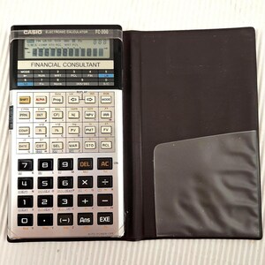 May include: A Casio FC-200 electronic financial consultant calculator with a silver face, black keys, and a digital display screen. The calculator is attached to a black folding protective case with a clear plastic pocket on the interior side.