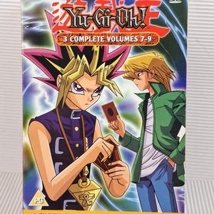 Puede incluir: Un set de DVD de Yu-Gi-Oh! que contiene 3 volúmenes completos 7-9. La portada muestra a dos personajes de anime sosteniendo cartas de juego sobre un fondo de rayos azules y morados. El texto dice 3 COMPLETE VOLUMES 7-9 y 9 EXCITING EPISODES.