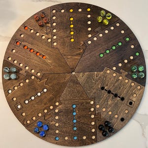 Könnte beinhalten: Ein kreisförmiges Holzbrettspiel, das in sechs Abschnitte unterteilt ist. Jeder Abschnitt hat einen anderen Satz farbiger Murmeln, darunter rot, gelb, grün, schwarz, blau und klar. Das Brett ist dunkelbraun gebeizt.