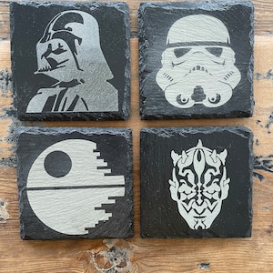 May include: Set of four black slate coasters with white images of Star Wars characters. The coasters feature Darth Vader, a Stormtrooper, the Death Star, and Darth Maul.