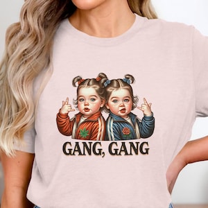 May include: A pale pink t-shirt with a graphic of two babies making rude gestures. The babies are in tracksuits, one red and one blue. The words "GANG, GANG" are printed beneath the image.