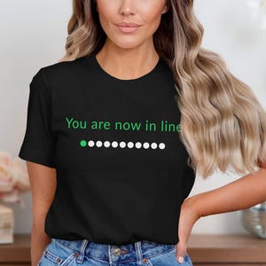 Puede incluir: Camiseta negra de cuello redondo con el texto "You are now in line" en verde y blanco. Una serie de puntos blancos sigue a un solo punto verde, lo que sugiere un estado de carga o espera. La camiseta la lleva una persona.