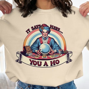 May include: A light beige sweatshirt with a graphic of a fortune teller with a crystal ball and tarot cards. The text on the sweatshirt reads "It says here... You a ho."