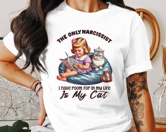 May include: White t-shirt with a graphic featuring a vintage illustration of a girl reading to a cat wearing a crown. Text reads "THE ONLY NARCISSIST I have room for in my life Is My Cat."