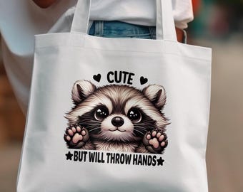 May include: White tote bag featuring a cartoon raccoon with the text "Cute but will throw hands" printed on it.