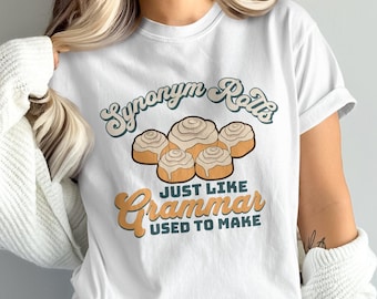 Puede incluir: Camiseta blanca con un diseño gráfico que presenta el texto "Synonym Rolls Just Like Grammar Used To Make". El gráfico también incluye una imagen de dibujos animados de cinco rollos de canela.