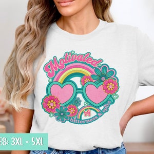 May include: White t-shirt with a graphic design of a rainbow, heart-shaped sunglasses, and flowers. The text "Motivated by bitterness & spite" is written below the sunglasses.