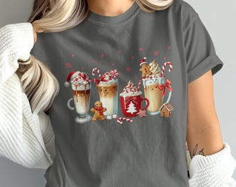 Puede incluir: Camiseta gris con una ilustración de acuarela de cinco bebidas festivas. Cada bebida está adornada con crema batida, bastones de caramelo y adornos de pan de jengibre. Copos de nieve rojos están dispersos en la parte superior.