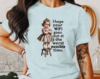 May include: Light blue t-shirt featuring a vintage-style illustration of an angry girl standing on a stool. The text on the shirt reads, "I hope your WiFi goes out at the worst possible time."