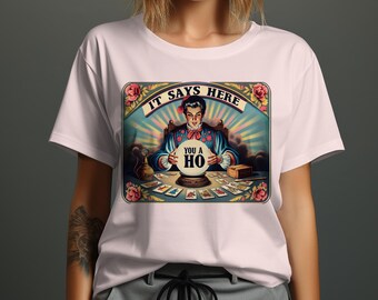 Puede incluir: Una camiseta rosa con una ilustración de estilo vintage de una adivina sosteniendo una bola de cristal que dice "You a HO". La ilustración está rodeada de un marco colorido con el texto "IT SAYS HERE" arriba.