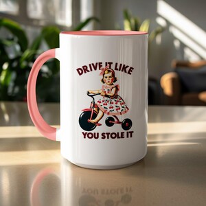 May include: White ceramic mug with a pink interior and handle. The mug features a vintage illustration of a girl on a tricycle with the text "DRIVE IT LIKE YOU STOLE IT" in maroon.