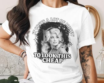 May include: White t-shirt featuring a black and white image of a woman with the text "IT COSTS A LOT OF MONEY TO LOOK THIS CHEAP". The graphic is surrounded by a black circle and stars.