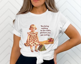 May include: White t-shirt with a graphic of a young girl sitting on a seesaw with strawberries. The text on the shirt reads "You bring nothing to the table, yet here you are eating."