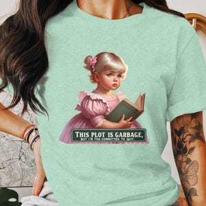 Może przedstawiać: Jasnozielony t-shirt z grafiką przedstawiającą młodą dziewczynę w różowej sukience trzymającą książkę. Tekst na koszulce brzmi "This plot is garbage, but I'm too committed to quit."