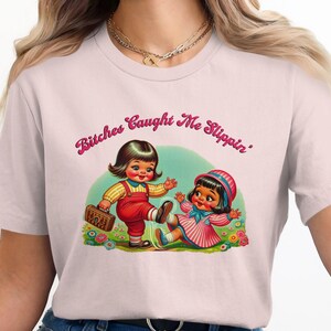 Peut inclure: T-shirt rose clair avec une illustration rétro de deux fillettes de dessin animé. L'une donne un coup de pied à l'autre, avec le texte "Bitches Caught Me Slippin'" en script rose au-dessus. Le design est sur fond bleu clair.