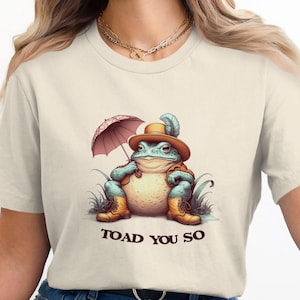 May include: A beige t-shirt featuring a cartoon illustration of a toad wearing a hat and holding an umbrella. The toad is sitting on a patch of grass. The text "TOAD YOU SO" is printed below the toad.