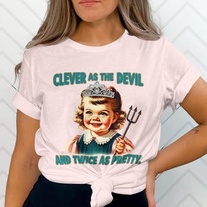 May include: Pink t-shirt featuring a graphic of a girl wearing a crown and holding a trident. The text on the shirt reads "Clever as the devil and twice as pretty."