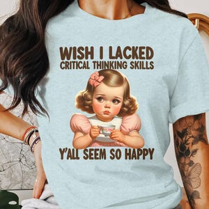 May include: A light blue t-shirt with a graphic of a sad girl holding a teacup. The text on the shirt reads "Wish I lacked critical thinking skills Y'all seem so happy."