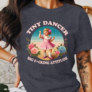 May include: Dark gray t-shirt featuring a graphic of a young girl in a pink dress, with the text "TINY DANCER" and "BIG F*CKING ATTITUDE" in a circular design. The design includes flowers and a striped background.