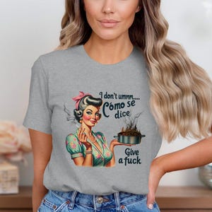 Peut inclure: Un t-shirt gris avec une illustration de style rétro d'une femme tenant une casserole avec des flammes. Le texte sur le t-shirt se lit "I don't ummm... Cómo se dice Give a fuck."