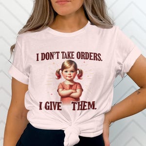 May include: A white t-shirt with a pink hue featuring a vintage-style illustration of a young girl with pigtails and the text "I don't take orders. I give them."