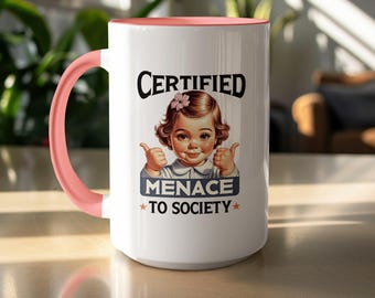 May include: White ceramic mug with a pink handle and interior. The mug features a vintage illustration of a child with the text "CERTIFIED MENACE TO SOCIETY". The design includes a pink flower and stars.