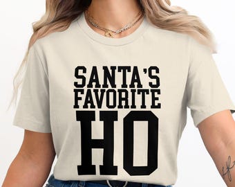 May include: Cream-colored t-shirt with the text "SANTA'S FAVORITE HO" in bold, black, collegiate-style lettering. The shirt is a casual, everyday wear item.