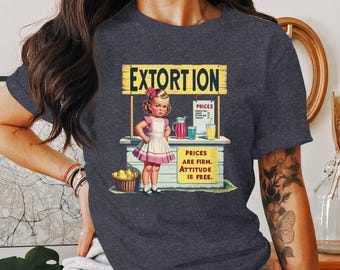 May include: A dark blue t-shirt with a vintage-style graphic of a girl selling lemonade. The graphic says "Extortion" and "Prices are firm. Attitude is free."