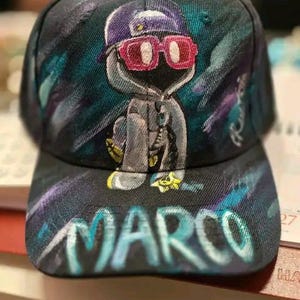 Può includere: Cappellino da baseball nero con un design dipinto personalizzato. Il cappellino presenta un personaggio dei cartoni animati con occhiali da sole e una felpa con cappuccio, con il nome "MARCO" a grandi lettere sul davanti. Il design incorpora sfumature di viola, verde acqua e rosa.