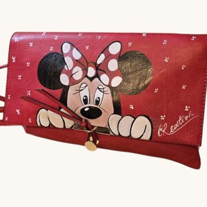 May include: Red leather clutch featuring a hand-painted Minnie Mouse design. The clutch has a flap closure with a gold-colored button and a shoulder strap. The Minnie Mouse design includes a polka-dot bow and the word "Creation" is written on the side.