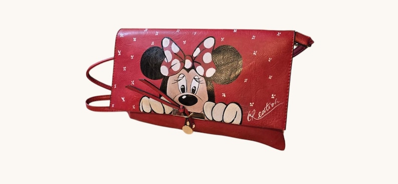 May include: Red leather clutch featuring a hand-painted Minnie Mouse design. The clutch has a flap closure with a gold-colored button and a shoulder strap. The Minnie Mouse design includes a polka-dot bow and the word "Creation" is written on the side.