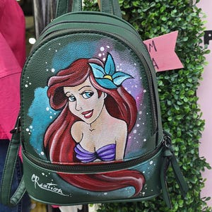 Puede incluir: Mochila verde con una imagen pintada a mano de Ariel de La Sirenita. El diseño incluye el pelo rojo de Ariel, ojos azules y una flor azul. La mochila tiene cierre de cremallera y correas ajustables.