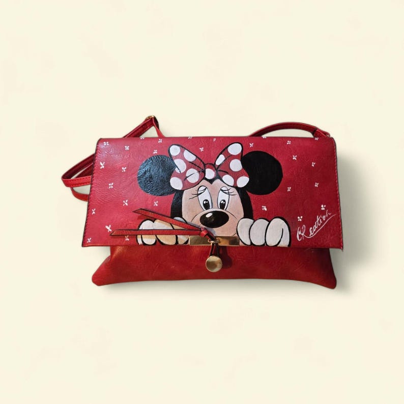 May include: Red crossbody bag featuring a hand-painted Minnie Mouse design. The bag has a flap closure with a gold-tone clasp and a long strap. The Minnie Mouse design includes a polka-dot bow and is set against a red background.