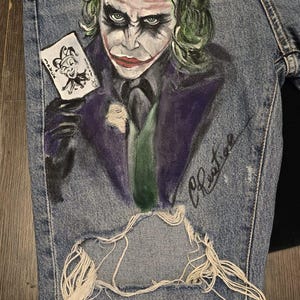 May include: Light blue denim jeans featuring a hand-painted image of the Joker holding a playing card. The Joker's face is painted with green hair, white face paint, and red lipstick. The jeans have a distressed, ripped knee.