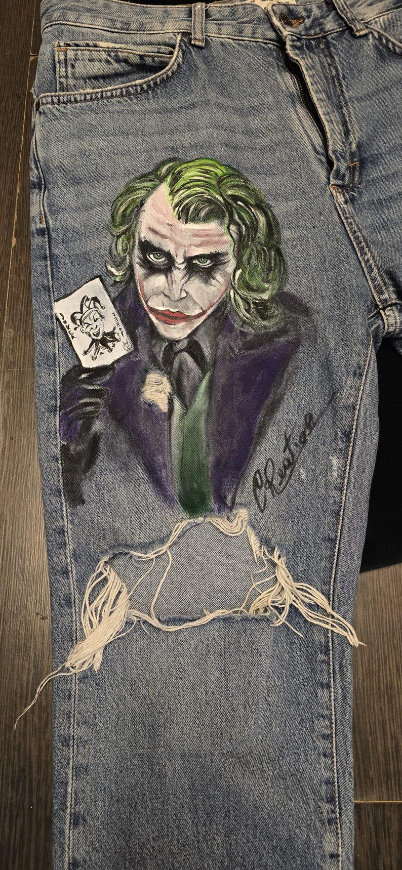 May include: Light blue denim jeans featuring a hand-painted image of the Joker holding a playing card. The Joker's face is painted with green hair, white face paint, and red lipstick. The jeans have a distressed, ripped knee.
