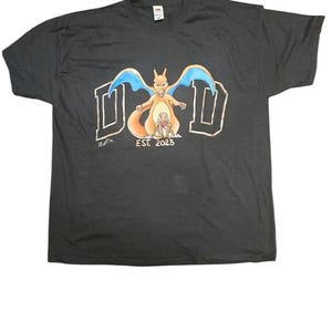 May include: Black t-shirt with a cartoon dragon graphic, blue wings, and a smaller dragon. The word "DAD" is in a stylized font above the dragons, with "EST. 2023" below.