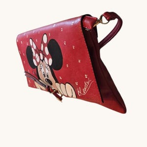 May include: Red handbag featuring Minnie Mouse with a white polka dot bow and a matching design. The bag has a shoulder strap and a clasp closure. The bag is a vibrant red color with a white signature.