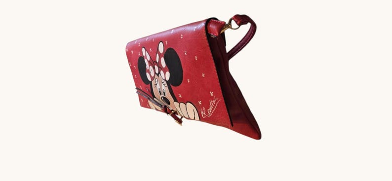 May include: Red handbag featuring Minnie Mouse with a white polka dot bow and a matching design. The bag has a shoulder strap and a clasp closure. The bag is a vibrant red color with a white signature.