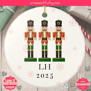 May include: A white ceramic Christmas ornament featuring three nutcracker soldiers in red, green, and black uniforms. The ornament includes the initials "LH" and the year "2025". The Etsy shop name "ornamentifi.etsy.com" is visible at the top.