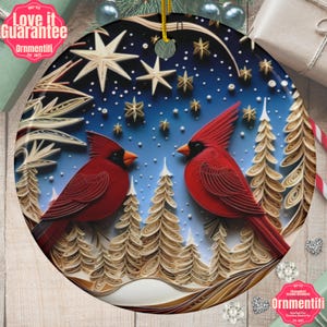 May include: A round Christmas ornament featuring a winter scene with two red cardinals perched on branches in a snowy forest. The background is a blue sky with white snowflakes and gold stars. The ornament has a white border and a gold string for hanging.