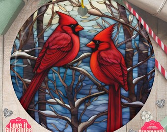 May include: A round ceramic ornament featuring a stained glass style illustration of two bright red cardinal birds perched on bare, snow-dusted tree branches against a blue and white winter sky. A gold string is attached to the top for hanging.
