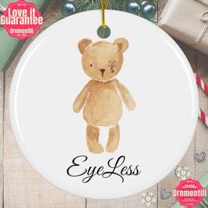 May include: A white ceramic ornament with a watercolor illustration of a brown teddy bear with a bandage over one eye. The text "Eye Less" is written in black cursive below the bear.